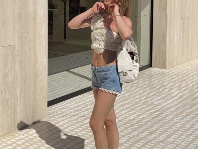 TheHottestBlonde has a small, fluffy white top, blue denim shorts with frayed edges, and a white bag, showing her slim, tan, and flat-assed body, with blonde hair.