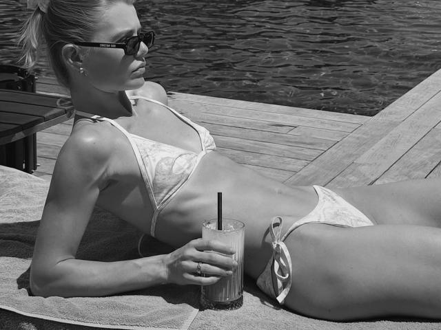 TheHottestBlonde, lying on a towel, wears a white bikini, showing her small tits and ass, holding a drink with a straw, wearing dark sunglasses, by a wooden deck.