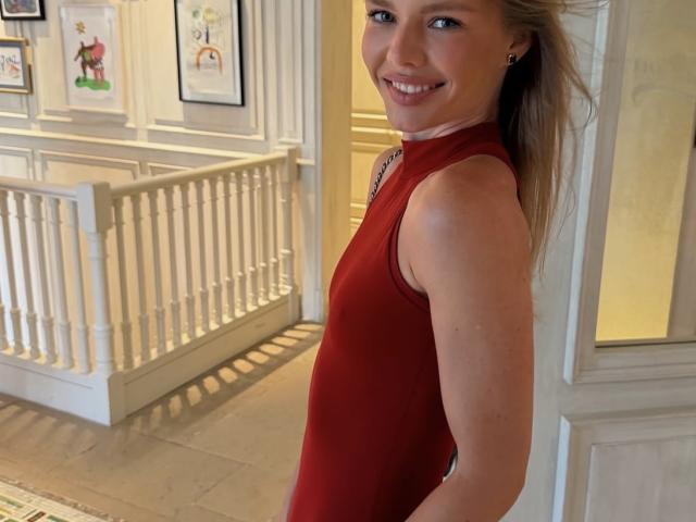 TheHottestBlonde, with a cute smile, wearing a red sleeveless top, showing her slim, attractive ass, and blonde hair in a ponytail, stands in a bright room.