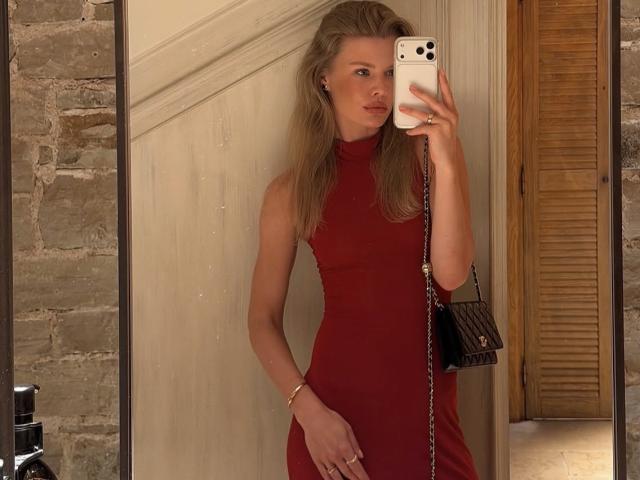 TheHottestBlonde with long blonde hair, red sleeveless dress, holding phone in front, small black purse on right, standing in front of mirror, looking at camera.