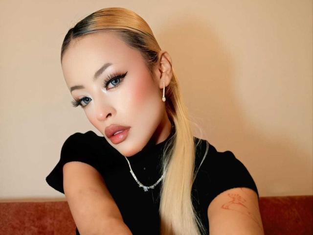 Angelique Hafner tem cabelo loiro, olhos azuis e batom rosa, vestindo uma blusa preta com um colar de prata. Ela tem uma tatuagem no braço direito..