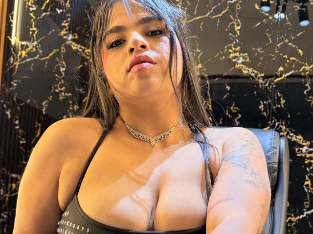Karina Mendez, com cabelo preto molhado, vestindo uma blusa cinza de decote baixo que mostra seus seios, um colar de prata, uma tatuagem no braço direito, olhando para a câmera com os lábios levemente entreabertos..