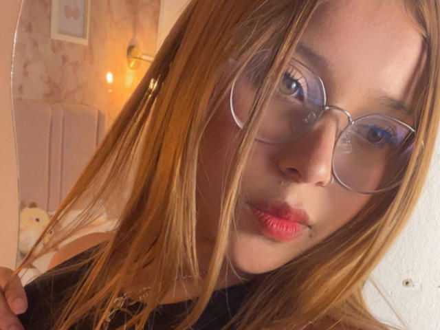 HeraJuno met rode lipstick, middelgrote borsten, een bril en lang blond haar, die een haarstreng vasthoudt, met een fronsende uitdrukking, in warm, binnenlicht..