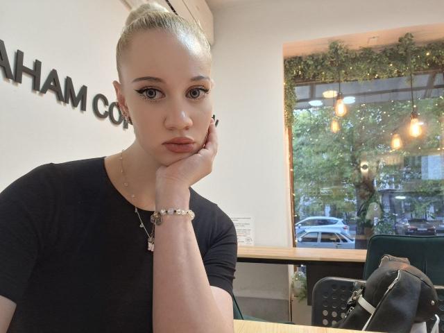 LindaxxCrow with blonde hair in a bun, black shirt, and wrist bracelet, resting chin on hand, looking at camera, in cafe, with black purse on table.