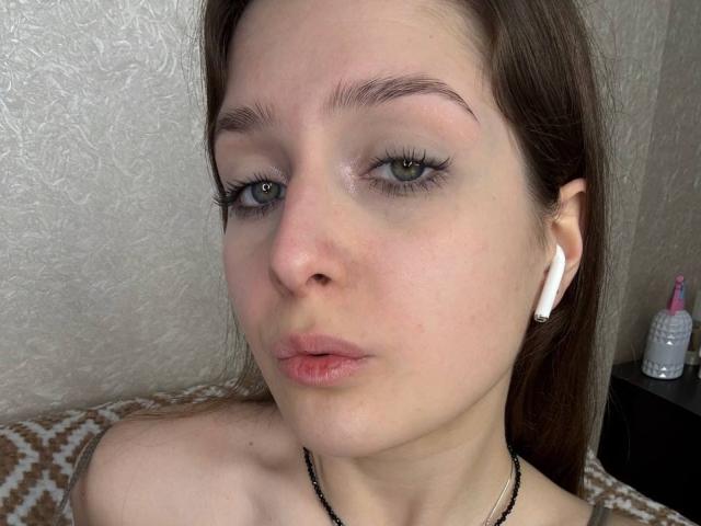 Sabina Grigalonis, con la pelle chiara, gli occhi marroni e i lunghi capelli castani, indossa auricolari bianchi, una collana nera ed è nuda. Ha le labbra rosa leggermente dischiuse, è corrucciata e guarda la fotocamera..