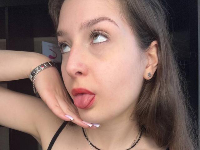Sabina Grigalonis tem pele clara, cabelo castanho e está de língua para fora, com batom vermelho, olhando para cima, vestindo uma blusa preta com alças e uma pulseira de prata..