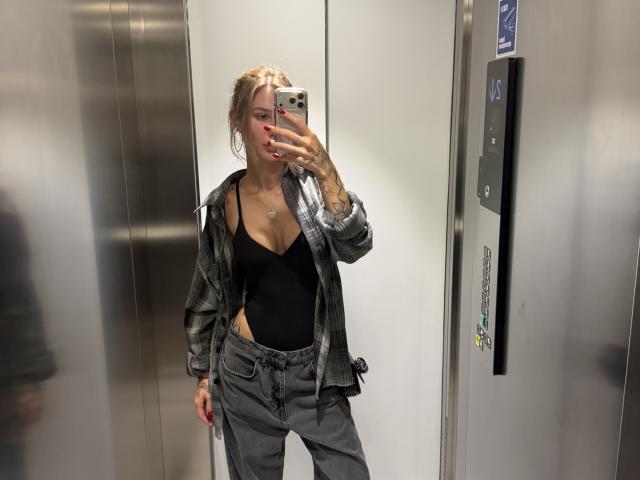 SensualMila, com cabelo loiro, regata preta, camisa xadrez cinza e calça cinza, tirando uma selfie no espelho dentro de um elevador, mostrando seus seios e o braço tatuado..
