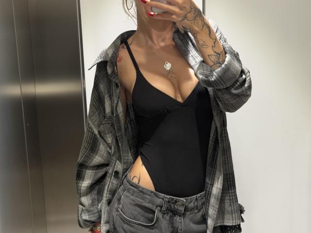 SensualMila, con tatuaggi, indossa una body nera a slip, una camicia a quadri e pantaloni grigi, tiene in mano un telefono, mostrando il seno e un tatuaggio sulla parte destra del fianco..