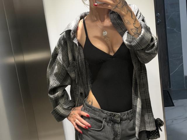 SensualMila with tattoos, wearing a black thong bodysuit, grey plaid shirt, and grey pants, showing her small tits and a heart necklace, taking a mirror selfie.