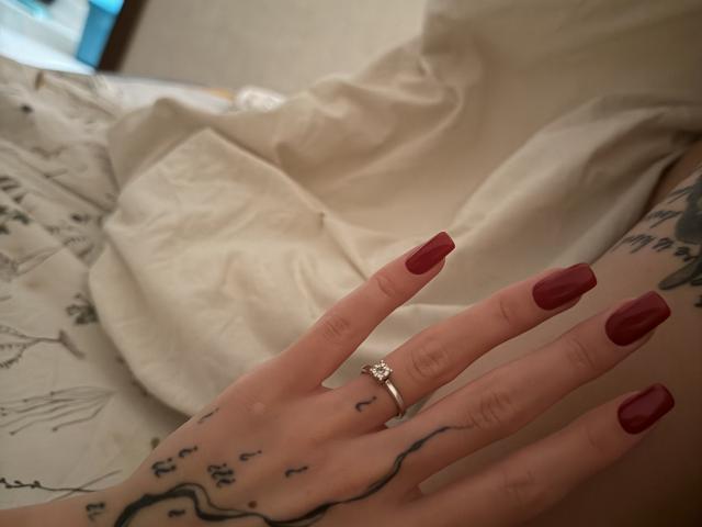 SensualMila's hand with red nail polish, a diamond ring, and tattoos on fingers and wrist, lying on white sheets. Boobless, assless, pussyless. Soft, intimate bedroom setting.