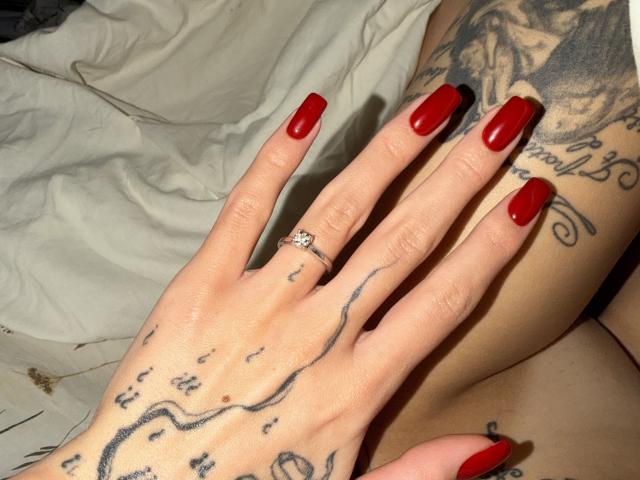 SensualMila's hand with red nails, a diamond ring, and black line tattoos, showing off her inked arm and leg. Her ass and tattoos are partially visible.