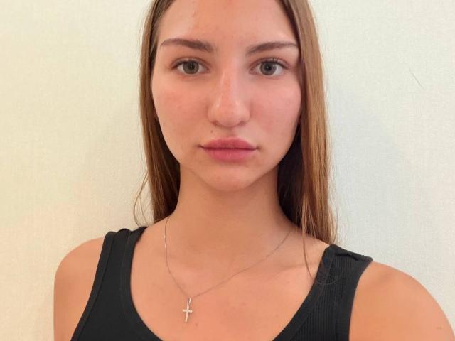 Adriana Nalley hat ein hübsches Gesicht mit grünen Augen, hellbraunen Haaren und einem kleinen Nasenpiercing. Sie trägt ein schwarzes Tanktop und eine Kreuzkette..