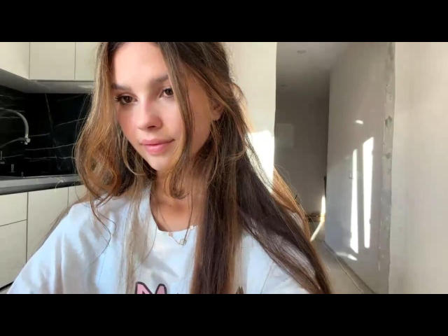 ElinaSoucie hat langes, braunes Haar, ein süßes Lächeln und kleine Brüste. Sie trägt ein weißes Hemd und steht in einer Küche, durch ein Fenster scheint Licht..
