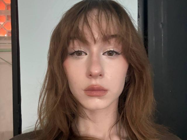 ClaudetteBemo a les cheveux bruns et en désordre, une peau pâle et des yeux bruns avec un trait d'eye-liner discret. Ses lèvres sont légèrement entrouvertes, laissant apparaître une expression neutre. Elle porte un haut noir..