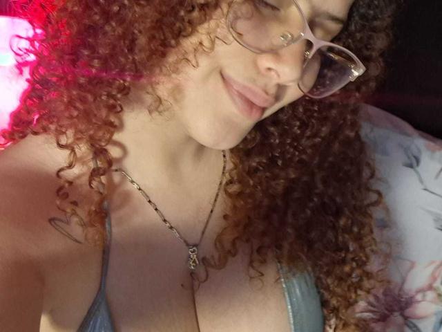 HoneyHipsss, con cabello rizado y rojo, gafas y una sonrisa, mostrando sus grandes senos en una blusa azul, llevando un collar con un tatuaje de corazón visible en su hombro derecho..