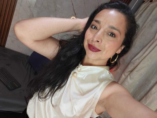 StangelarRivera has long black hair, red lipstick, hoop earrings, and a shiny white sleeveless top, with a hand behind her head, showing armpit hair.