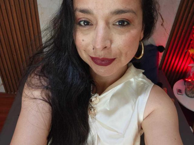 StangelarRivera with long black hair, wearing a white satin sleeveless top, gold hoop earrings, and dark red lipstick, smiling at the camera, showing a small mole on her left cheek.