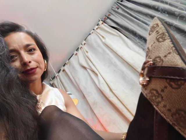 StangelarRivera, with long black hair and dark red lipstick, wears a white top and black stockings, looking down at a brown and beige Gucci loafers.