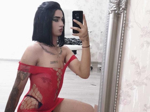 IrisIrs with black hair, red fishnet lingerie, tattoos, holding phone, mirror selfie, showing small tits, flat ass, and slim build, kneeling on wooden floor, looking at phone.
