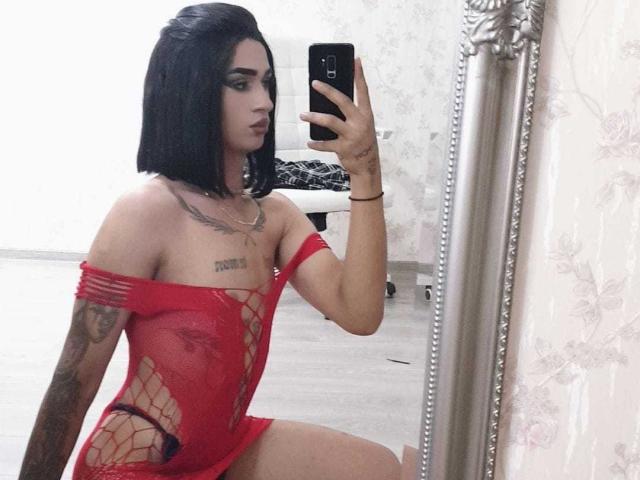 IrisIrs with black bob, red fishnet dress, tattoos, holding phone in mirror selfie, showing off small tits and tattooed arms, kneeling on wooden floor.
