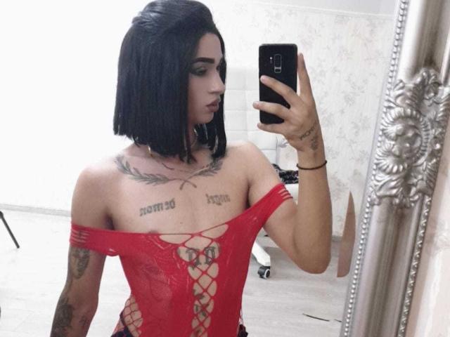 Iris, avec une coupe de cheveux courte noire, un body rouge à lacets, des tatouages, tenant un téléphone pour un selfie dans un miroir, montrant de petites seins, des tatouages sur la poitrine et des tatouages sur les bras, dans une pièce avec des miroirs..