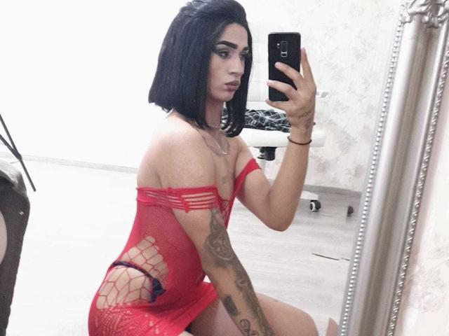 Iris, avec une coupe de cheveux courte noire, une robe en filet rouge, montrant des tatouages, tenant un téléphone, prenant un selfie devant un miroir, révélant une culotte rouge, agenouillée, regardant le téléphone, avec des seins de taille moyenne et un joli visage..