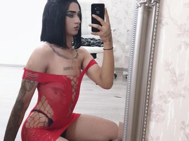 IrisIrs with black bob, wearing red fishnet dress, sitting, showing tattoos, holding phone, mirror selfie, visible breasts, nipples through mesh, arm tattoo, leg tattoo, room.