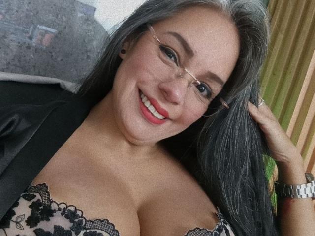 LailaCastillo with gray hair, glasses, and red lipstick, smiling at the camera, showing her large boobs in a lace bra, wearing a black jacket, hand in hair.