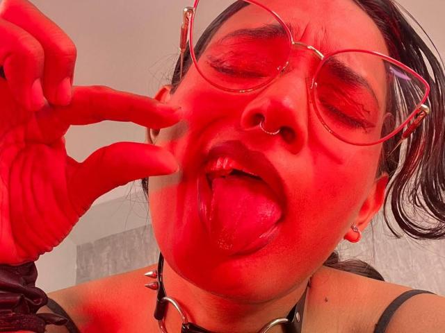 LilithRussoo with red tinted skin, tongue out, wearing red glasses and a black collar with metal rings. She's making a peace sign near her face, eyes closed.