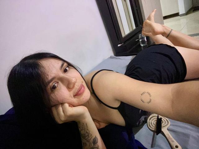 SophiaMarx with black hair, lying on a bed, black tank top, black shorts, tattoo on right forearm, smiling, head on hand, feet up, mirror in background.