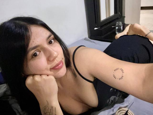 SophiaMarx with black hair, medium tits, black tank top, lying on bed, arms tattooed, smiling, head resting on hand, black frame mirror in background, water bottle.