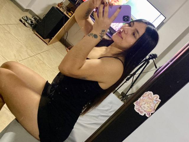 SophiaMarx with long black hair, small tattoo on right wrist, wearing a black dress, taking a mirror selfie, showing her ass, in a room with a bed and tripod.
