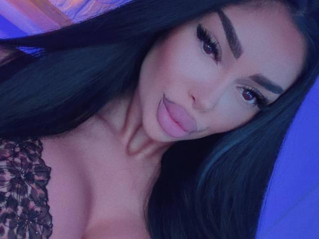 BlackClover with long black hair, thick eyebrows, and full pink lips, sticking out her tongue. She's wearing a lace top, showing a hint of her tits. Blue-lit face.
