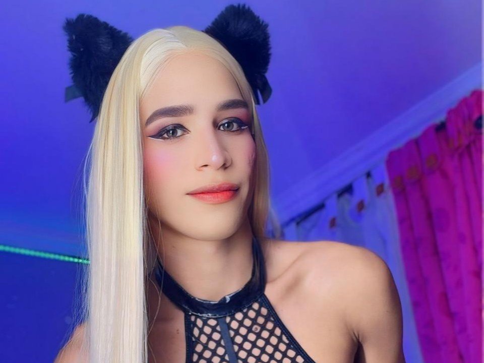 KingSamira, com cabelo loiro, orelhas de gato pretas e peludas, e uma blusa de meia-calça, exibindo seus seios, maquiagem rosa e batom laranja, sorrindo em um quarto com iluminação azul..