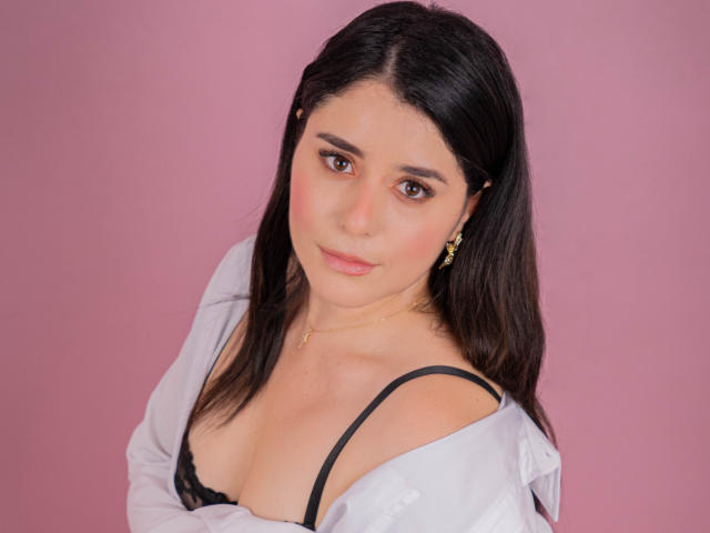 VickyMegan with dark hair, brown eyes, and a slight smile, wearing a black bra with a white shirt off her shoulders, showing her tits, against a pink background.