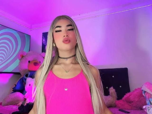 Heidy Klum mit langen blonden Haaren, einem pinken Tanktop, einer schwarzen Halskette und vollen Lippen, steht in einem Raum mit violetter Beleuchtung, umgeben von Stofftieren. Ihre Brüste sind klein..