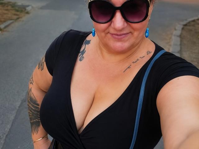 BlondyBigBoobs, con grandes senos, vestida con un vestido negro, aretes azules, gafas de sol negras, tatuajes en el hombro y el pecho, de pie en la calle, sonriendo, con una correa azul sobre el hombro..