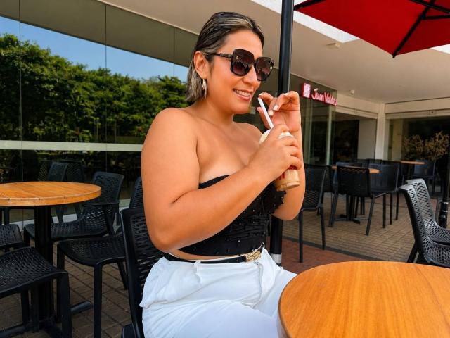 EmaFoxy with tan skin, dark brown hair, and sunglasses, sipping a frappuccino, wearing a black strapless top, white pants, and a gold belt, sitting at a wooden table.