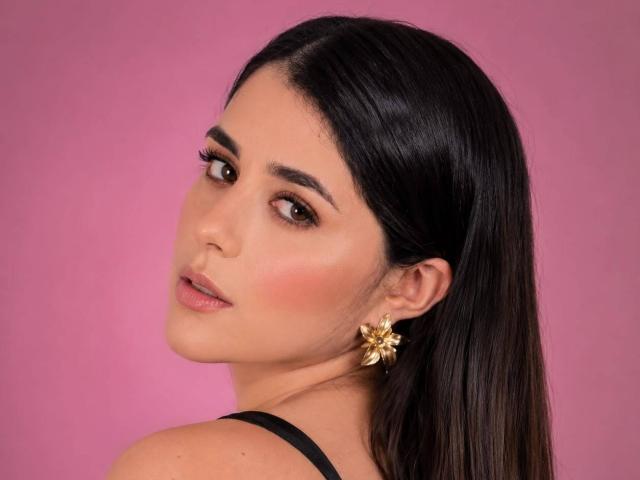 VickyMegan has dark, straight hair, brown eyes, and a pink blush. She's wearing a black strap and gold flower earrings, looking over her shoulder with a slight smile.