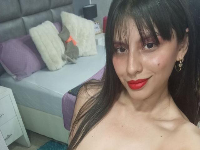 MadissonFontaine has long black hair, red lipstick, and is topless with small tits, wearing gold earrings, smiling at the camera in a bedroom with a gray headboard.