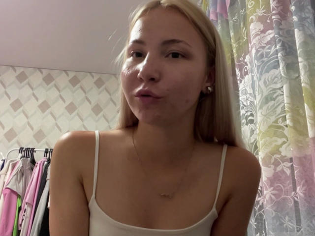 Marguerite Geppert ha i capelli biondo chiaro, la pelle chiara e piccoli seni, indossa una canottiera bianca, ha un'espressione accigliata con un leggero sorriso, porta orecchini di perle, in una stanza con una luce soffusa..