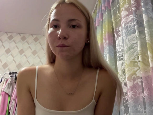 Marguerite Geppert mit blonden Haaren, einem weißen Tanktop und geschwollenen Lippen, trägt eine Halskette und befindet sich in einem Raum mit bunten, blumigen Vorhängen und einer gemusterten Wand. Ihre Brüste sind klein..