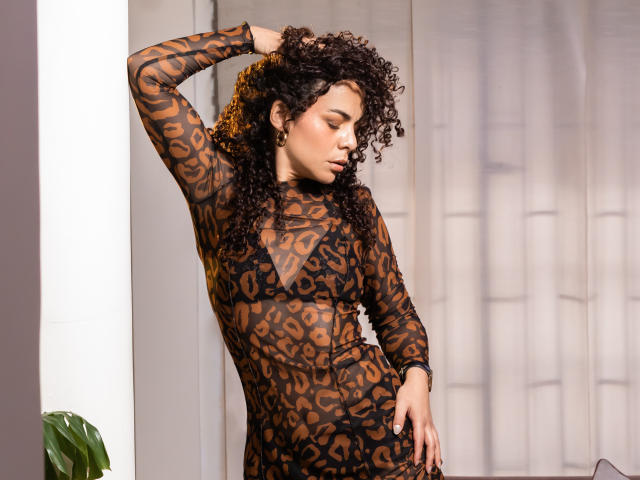MissSunshinee in a sheer, leopard-print dress, standing with one hand in curly brown hair, showing off her medium brown tits and smooth, brown skin. She's looking down, eyes closed.
