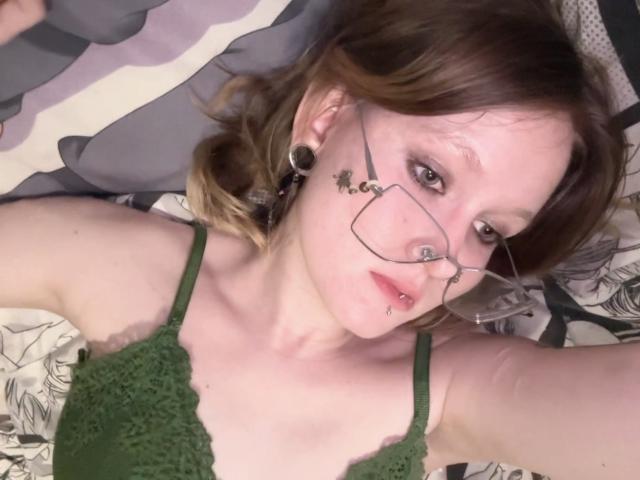 MozellContos with glasses, green lace top, pierced nose, black ear plugs, brown hair, lying on bed, looking at camera, fair skin, small tits, subtle makeup, relaxed expression.