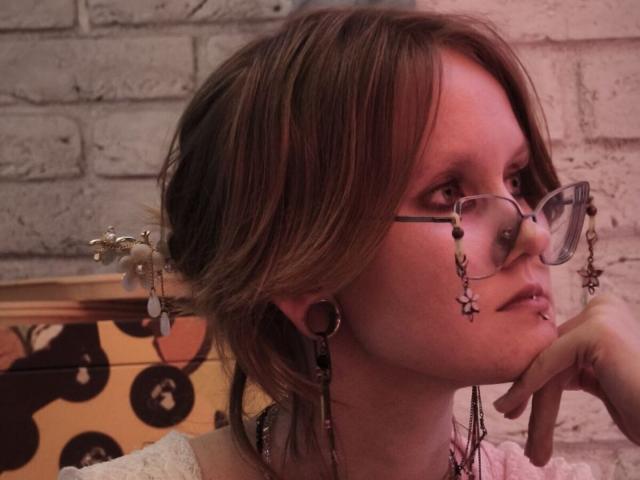 MozellContos with glasses, nose ring, and lip ring, wearing earrings and a pink top, resting chin on hand, brown hair, white brick wall, and wooden headboard in background.