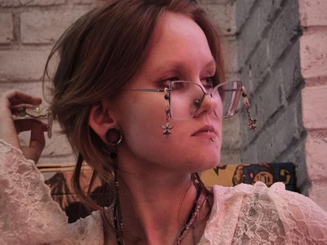 MozellContos with glasses, brown hair, and a nose ring, holding a glass, wearing a lace top, and large earrings, looking to the side with a slight pout.