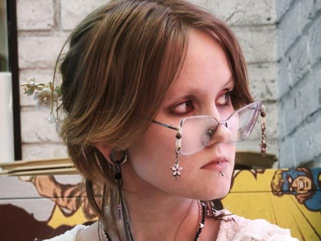 MozellContos has brown hair in a messy bun, wearing clear glasses with bead decorations, nose piercing, and lip ring, with large black earrings, against a white brick wall.