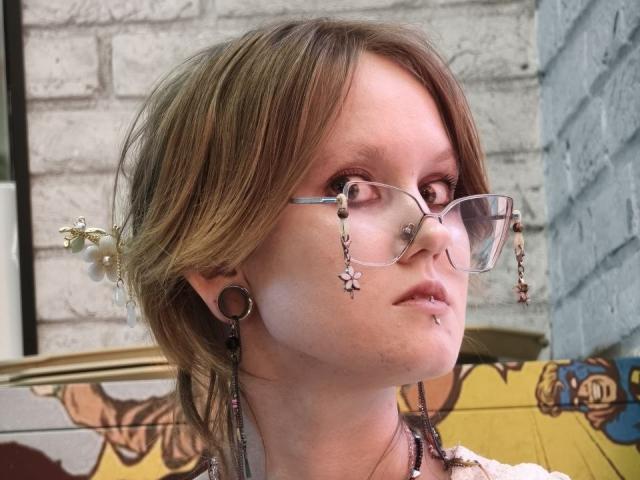 MozellContos has light brown hair, glasses with star charms, large ear gauges, and a nose piercing. She's looking up with a slightly pouty expression.
