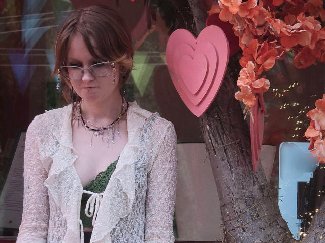 MozellContos with brown hair, glasses, white lace cardigan, green dress, and necklace, standing next to a tree with pink heart and orange flower decorations.
