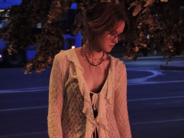 MozellContos has brown hair, wearing a lace, cream-colored, boho-style top with ruffles, and a gold necklace, standing under tree branches at night, looking down with a pensive expression.